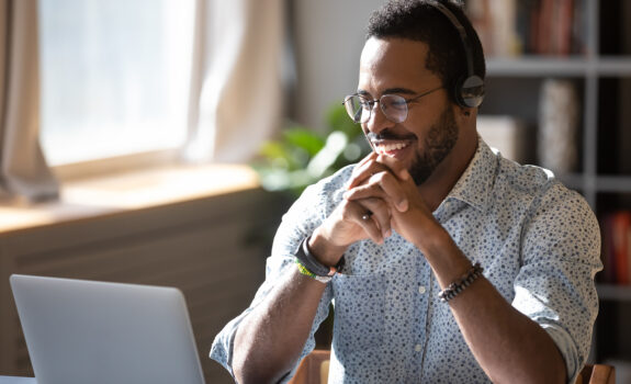 Happy millennial african american man in glasses wearing headphones, enjoying watching educational webinar on laptop. Smiling young mixed race businessman holding video call with clients partners.